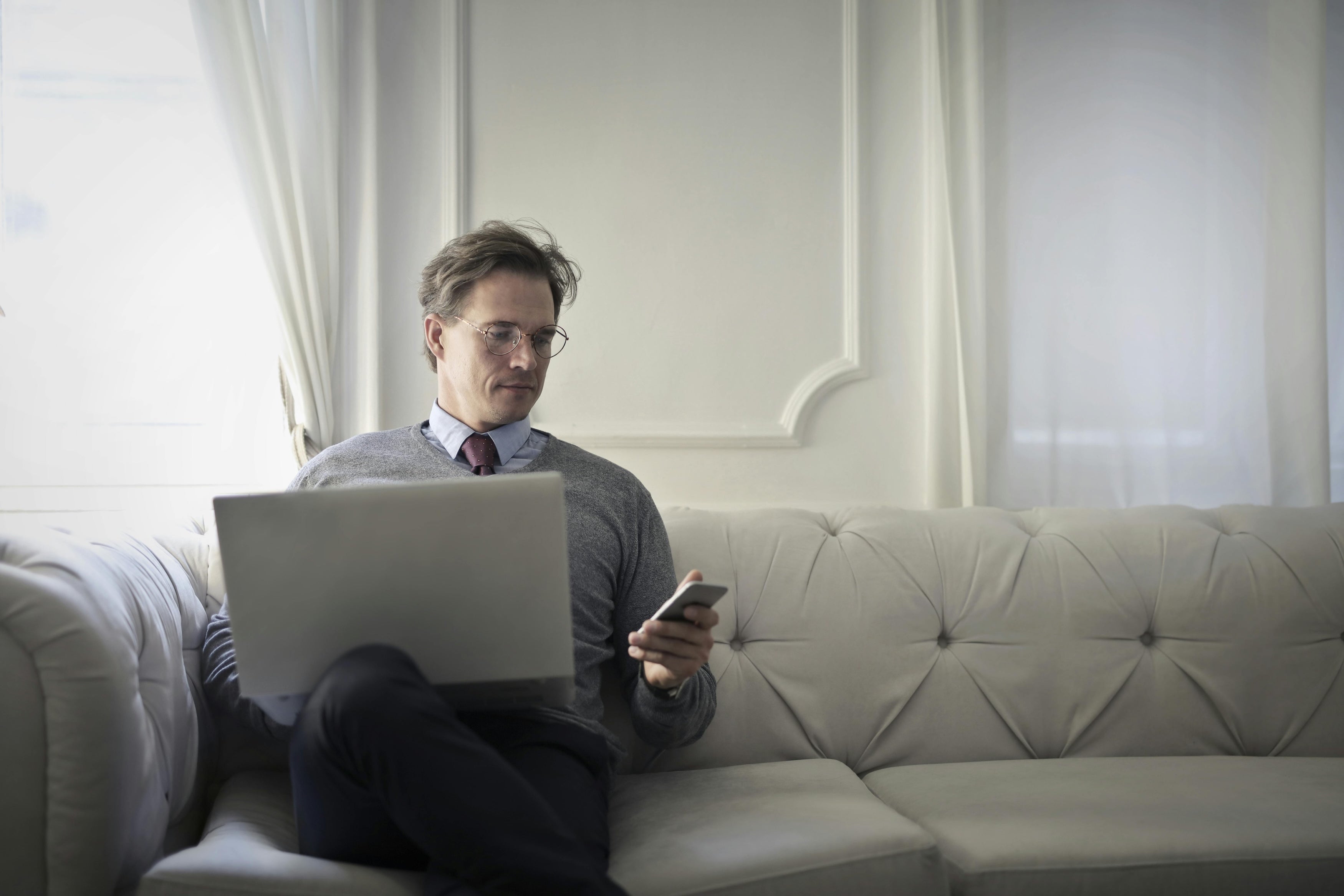 Affluent man in glasses sits on expensive couch, managing catalog of assets on computer and phone simultaneously.