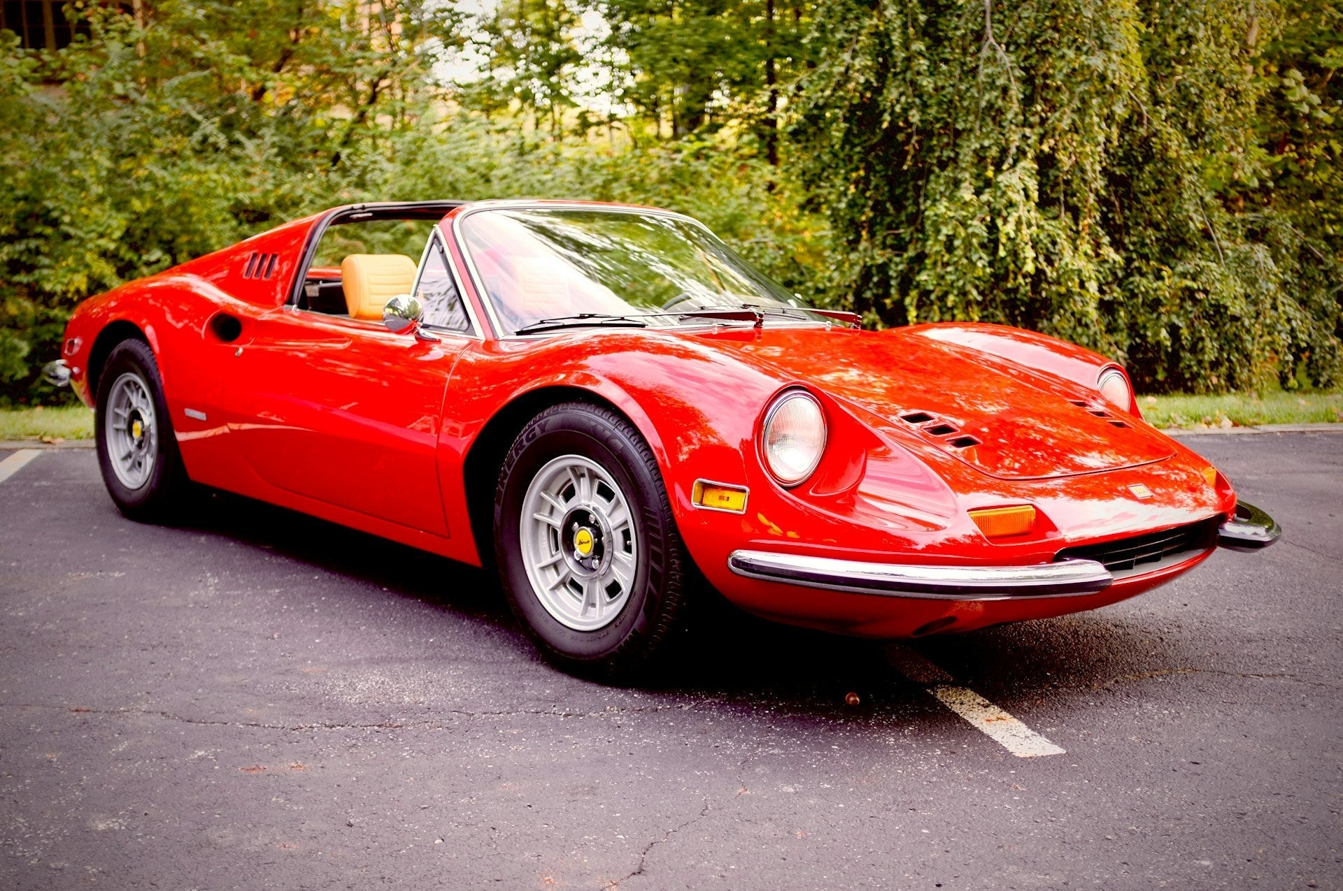 Classic red vintage sports car with sleek curves and chrome accents, parked on asphalt before a backdrop of lush green trees, tan leather interior visible through the open top.