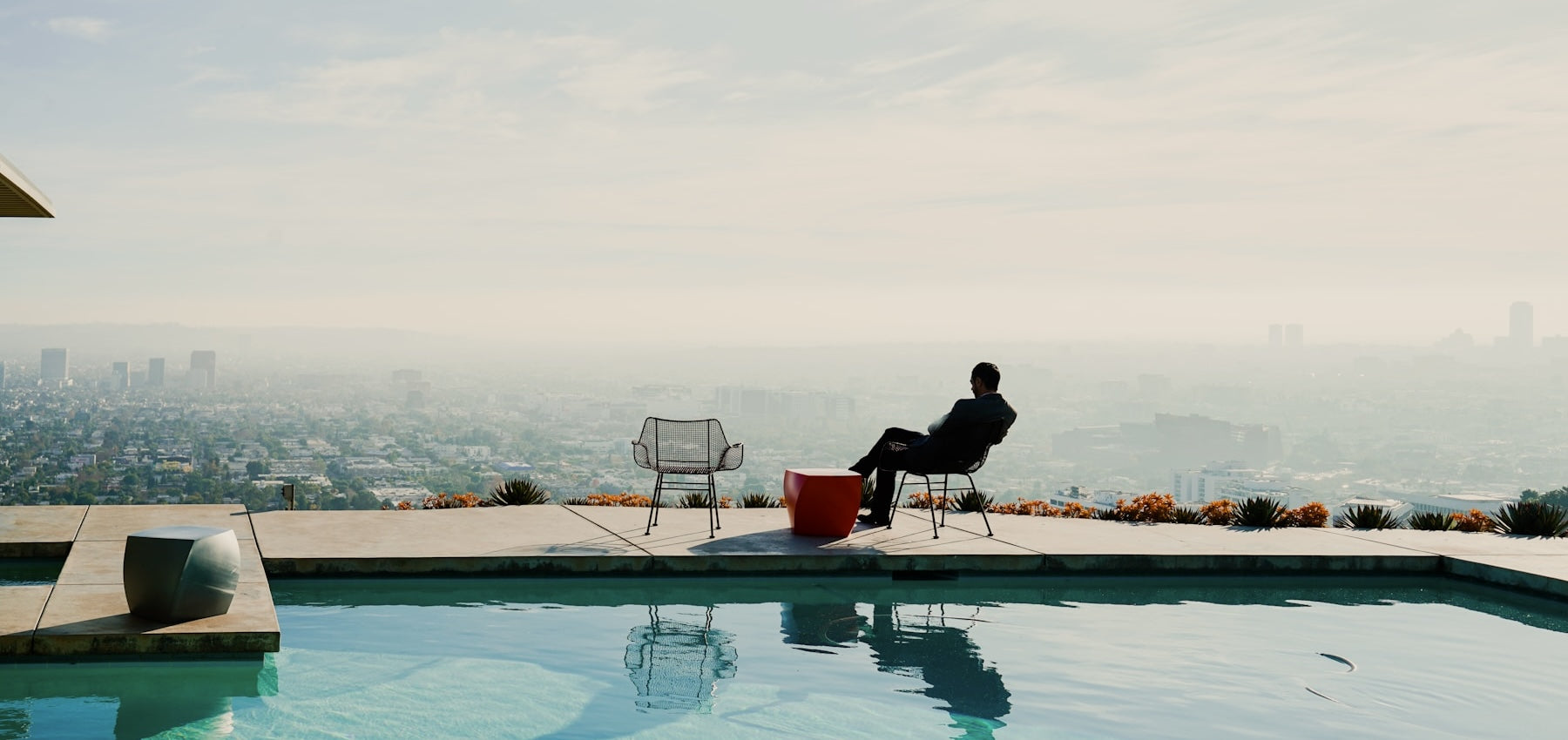 Man in a suit sits beside a rooftop pool, gazing over a hazy LA city skyline with high-rises in the distance, framed by minimal modern furniture and desert landscaping.