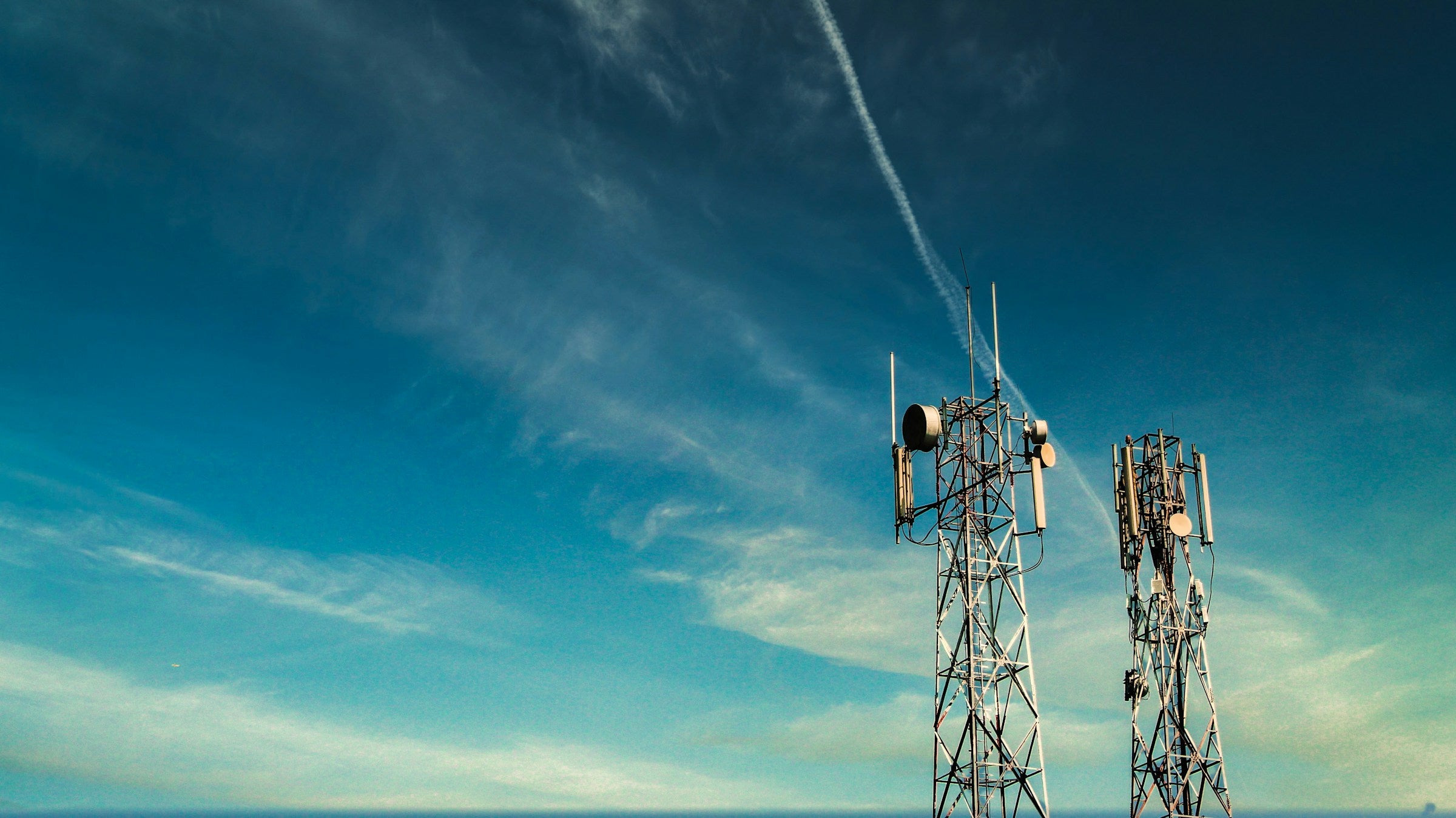Two tall cellular towers stand against a clear blue sky with wispy clouds and a visible contrail from an airplane.