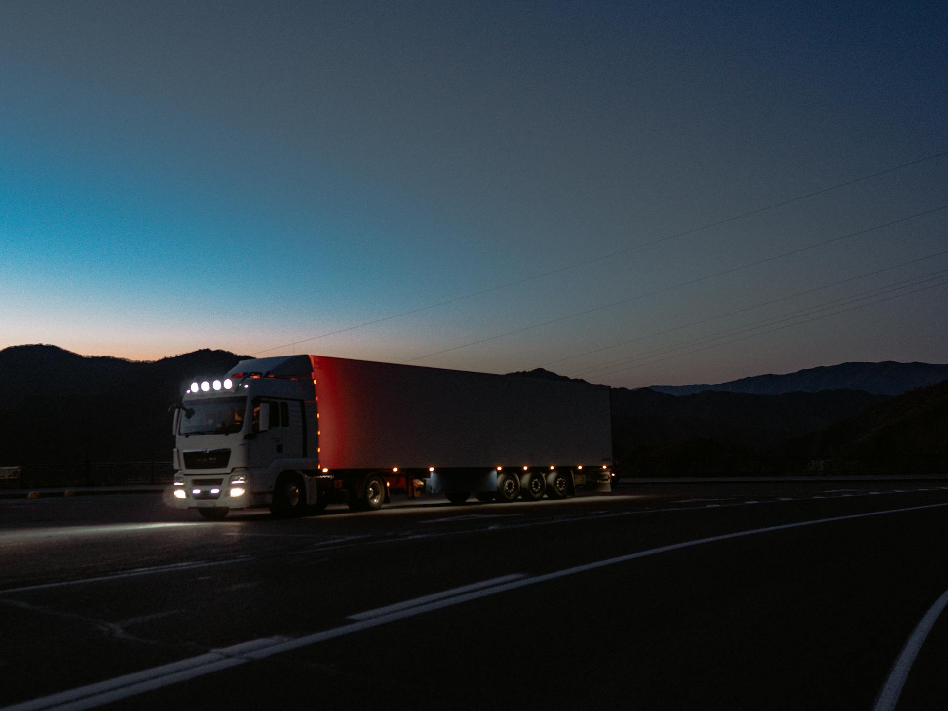 Long-haul transport truck illuminated in early evening light, driving on a curved highway through mountainous terrain, suggesting vehicle shipping or logistics risk for collectors